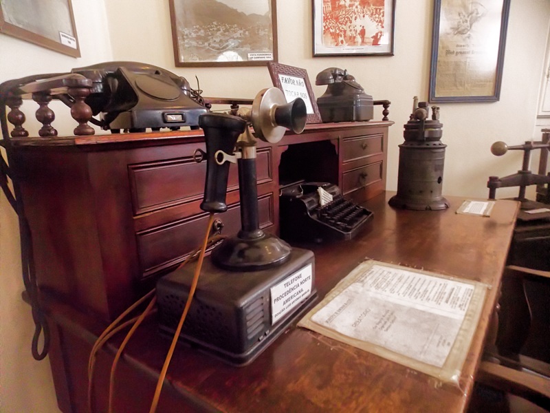 foto-demonstra-uma-antiga-escrivaninha-e-diversos-objetos-em-cima-como-um-antigo-telefone-e-um-telegrafo